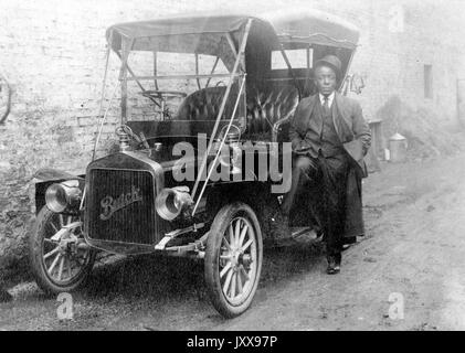 Full length shot of African American man leaning against a Buick car parked against a building, wearing a suit and hat tilted on his head, neutral facial expression, 1920. Stock Photo