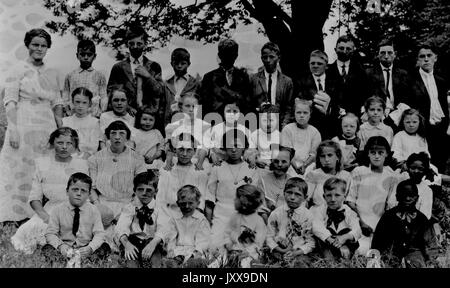 Landscape shot of schoolchildren, all seated in grass under a tree, one row standing, all boys and girls wearing white school uniforms, one African American boy in the bottom right row wearing a black shirt, neutral facial expressions, 1920. Stock Photo