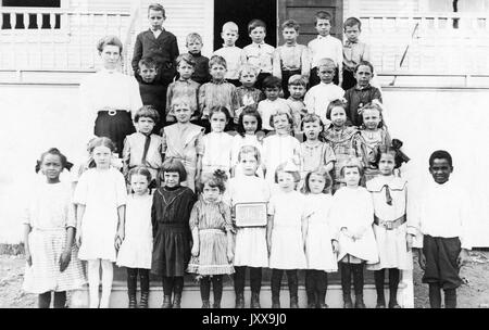 Full length portrait of Caucasian schoolchildren, all wearing uniforms, smiling facial expressions; two African American children on either side of the front row, 1920. Stock Photo