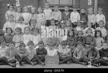 Full length landscape shot of schoolchildren, all seated except back row, one African American boy in the front row, holding a sign with teacher's name and room number, 1915. Stock Photo