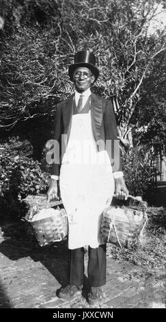 Full length standing portrait of a mature African American man with a neutral expression on a brick pathway in front of shrubbery, carrying a basket in either hand, wearing a top hat, glasses, a suit, a tie, and an apron, 1915.