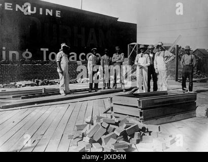 A group of American construction workers with neutral expressions stands outside of a tobacco company's building on a platform, with piles of bricks or pavers in front of and behind them, 1915. Stock Photo