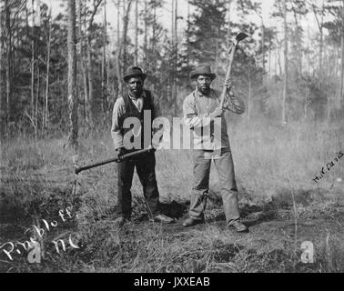 Full length standing portrait of two African American workers with neutral expressions, wearing hats, pants, shoes, button-up shirts and one wearing a suit vest, stand in a cleared wooded area with axes, posed as if they are at work, 1920. Stock Photo