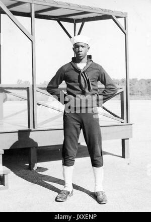 Portrait of an African American US Navy Sailor standing in front of a wooden platform, he is wearing a dark colored Sailor uniform and a light colored Sailor hat, his arms are bent and his hands are behind his back, 1920. Stock Photo