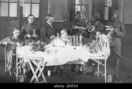 One white young man is perched up while his body lays on a long bed, there are other young white men sitting and standing near him holding knitting supplies, three African American men are standing behind the young man and are holding knitting supplies, a woman is instructing one of the young white men on what to do, 1920. Stock Photo
