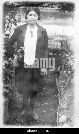 Portrait of an African American woman standing in front of a body of water, she is standing on dirt and to the left is shrubbery, she is wearing a dark colored knee-length skirt, a light color blouse and a dark long coat on top, her hair is separated into two low buns, 1920. Stock Photo