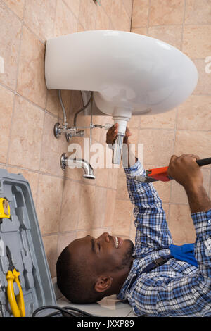 Young African Male Plumber Lying On Floor Fixing Sink In Bathroom Stock Photo