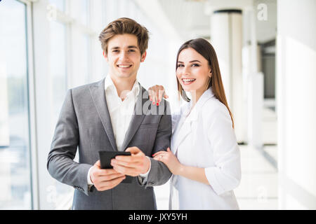 Two smiling elegant business people man in formal suit and woman in jacket and dress looking on digital tablet in office interior near window Stock Photo