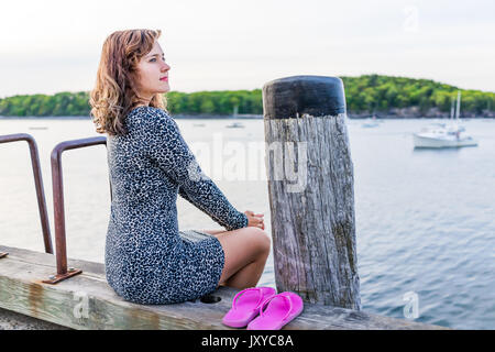 Young happy smiling woman sitting on edge of dock in Bar Harbor, Maine looking at water and bay Stock Photo