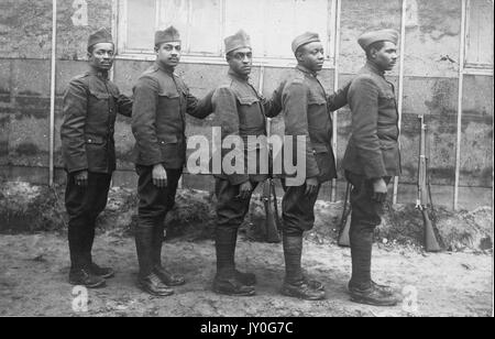 Standing group portrait of five African American soldiers with neutral expressions in uniform outside of a building, their weapons resting against its side, 1920. Stock Photo