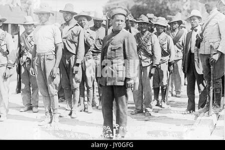 Young and mature African American soldiers with neutral expressions, some of whom carry weaponry, stand in rows outdoors, 1915. Stock Photo