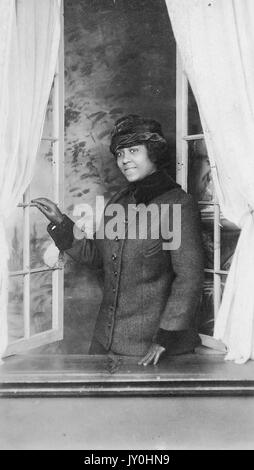 Half length standing portrait of young African American woman, wearing dark overcoat with hat, standing at windowsill while looking outside of open window, smiling expression, 1925. Stock Photo