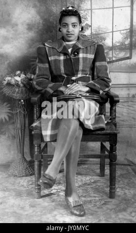 Full-body portrait of an African American woman sitting in a wooden chair, with her hands folded on her lap, legs crossed, wearing a large dark coat, seated in front of a backdrop, with a blank facial expression, 1920. Stock Photo