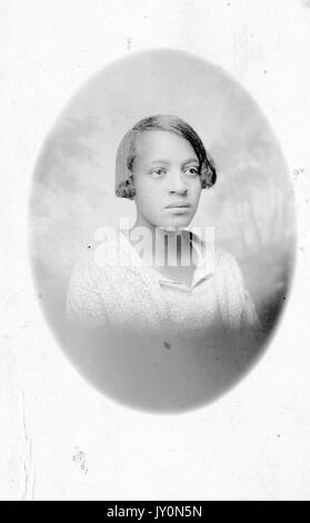 Oval head shot portrait of an African American woman, wearing a white blouse, with a serious facial expression, in front of a backdrop, 1920. Stock Photo