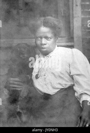 Three-quarter portrait of an African American Woman in front of a brick wall and window, wearing a white blouse and a dark dress, wearing a pendant around her neck, with a serious facial expression, 1920. Stock Photo