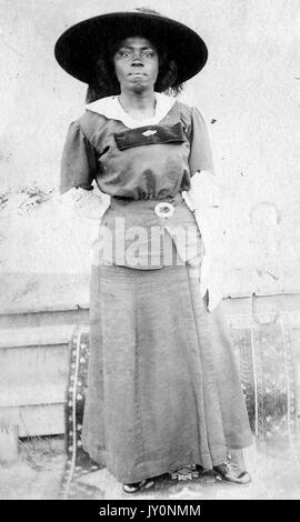 Full-body portrait of African American woman, wearing a dark dress and a large dark hat and white gloves, standing in front of a backdrop, with a serious facial expression, 1920. Stock Photo