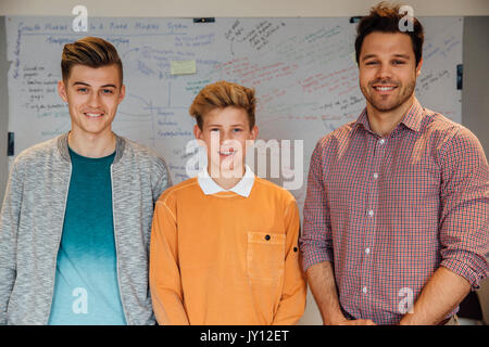Male teacher and two of his teen students are smiling for the camera in front of the whiteboard in a school classroom. Stock Photo