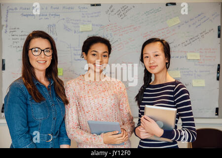 Female teacher and two of her teen students are smiling for the camera in front of the whiteboard in a school classroom. The students are holding digi Stock Photo