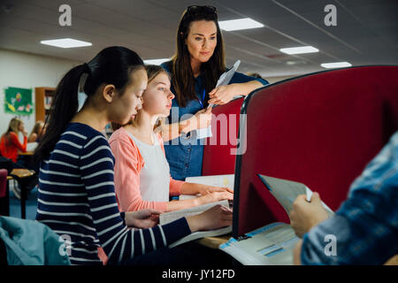 Students are working on computers with textbooks. A female teacher is standing with them, helping them with their work. Stock Photo
