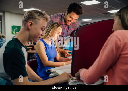 Students are working on computers with textbooks. A male teacher is standing with them, helping them with their work. Stock Photo