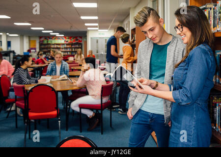 Female teacher is in the school library with her students. She is standing with one of her teen students and they are looking at a book together. Stock Photo