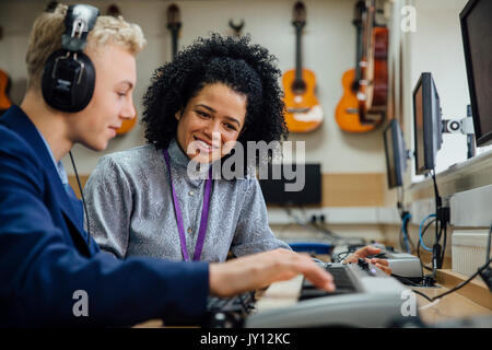 Female teacher is sitting with one of her students in a music lesson at school. He is learning to play the keyboard and is wearing headphones. Stock Photo