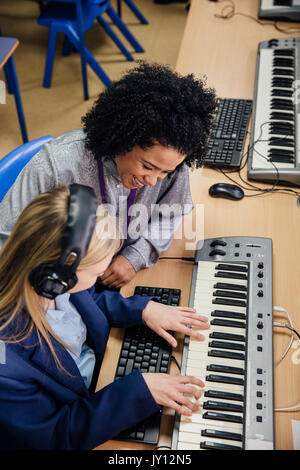 Female teacher is sitting with one of her students in a music lesson at school. She is learning to play the keyboard and is wearing headphones. Stock Photo