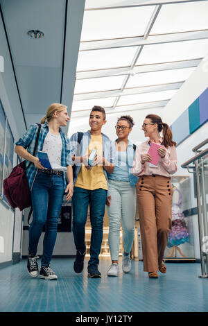 Teen students are walking down the school hall with their teacher. They are carrying books and talking. Stock Photo