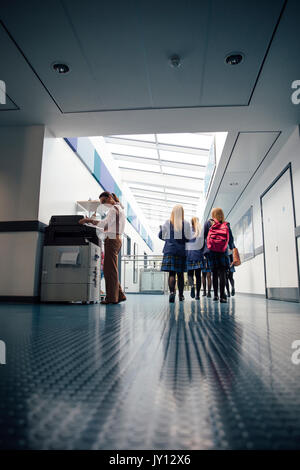 Group of students are walking down the corridor of their school. There is a female teacher using the printer. Stock Photo