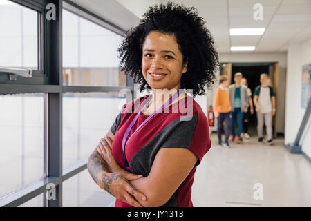 Portrait of a mixed race teacher with tattoos in the school hall. She has her arms folded and is smiling for the camera. Stock Photo