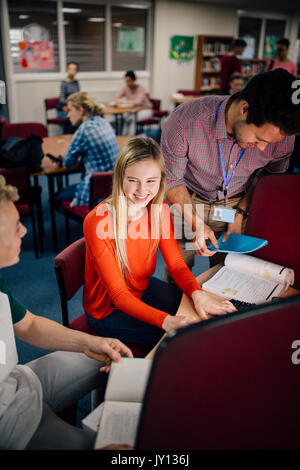Students are laughing as they work on computers together. There is a teacher leaning over their shoulder, helping them with their work. Stock Photo