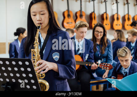 Teen student is playing the saxophone in her school music lesson. The rest of the class are in the background, out of focus. Stock Photo