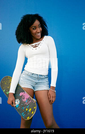 Smiling Black woman standing near blue wall holding skateboard Stock Photo