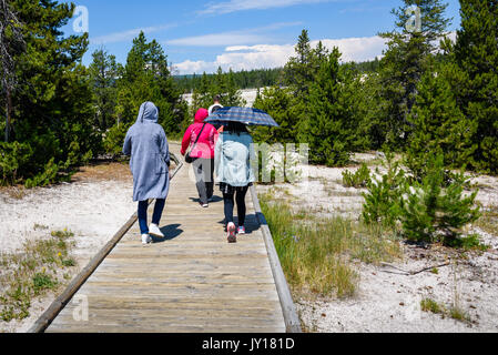 Tourists shaded by an umbrella and hoods walking in the sun on boardwalk in Yellowstone National Park, Wyoming USA Stock Photo