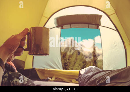 Man lying in tent with a coffee, views of the mountains and the sky from inside the tent. Camper in a sleeping bag looking at the beautiful scenery in Stock Photo