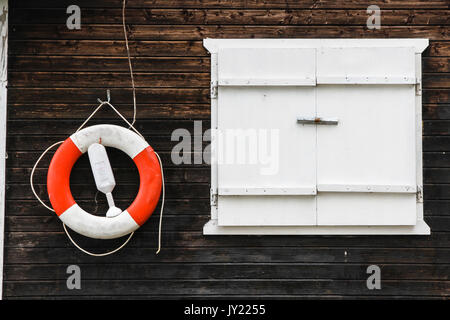 Red and white lifebuoy lifebelt with ropes hanging from a dark brown painted wooden wall with a white window on the right. Stock Photo