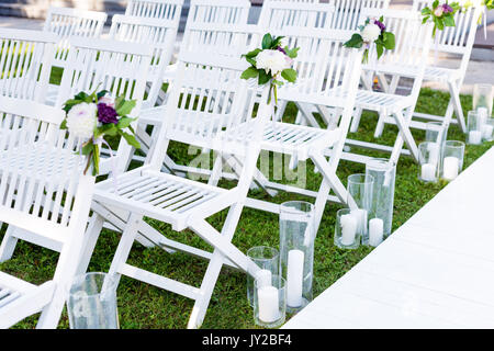 Beautiful wedding set up. Wedding ceremony in the garden. White wooden chairs decorated with flowers and candles standing in rows. Stock Photo