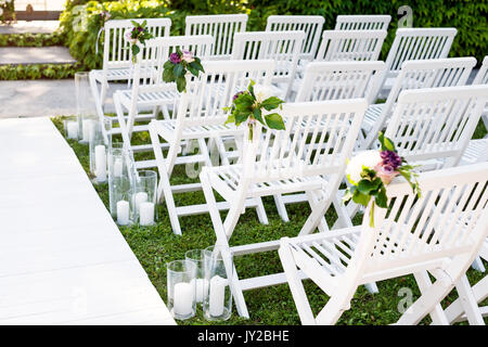 Beautiful wedding set up. Wedding ceremony in the garden. White wooden chairs decorated with flowers and candles standing in rows. Stock Photo