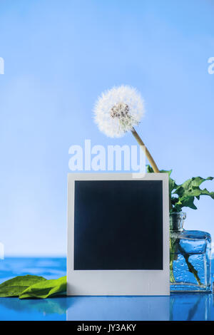 Simple still life with instant photo frame, summer rain, drops on a window and dandelions is a glass vase on a sky blue background. Copy space. Stock Photo