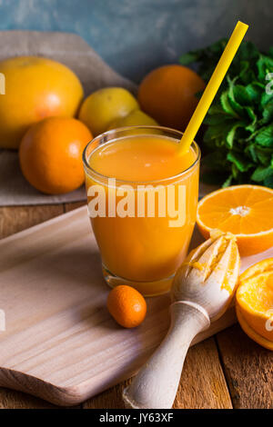 Freshly pressed citrus fruit juice from oranges lemons limes cumquat, glass with straw, reamer, mint on table by window, rustic kitchen interior Stock Photo