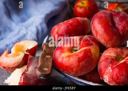 Bunch of ripe organic colorful red saturn peaches on white plate, halved, knife, blue napkin, dark kitchen table, rustic interior Stock Photo