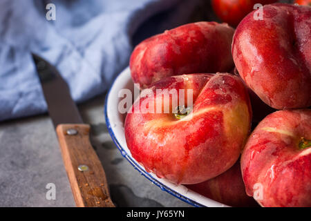 Bunch of ripe organic colorful red saturn peaches with water drops on white plate, knife, blue napkin, dark kitchen table, rustic interior Stock Photo