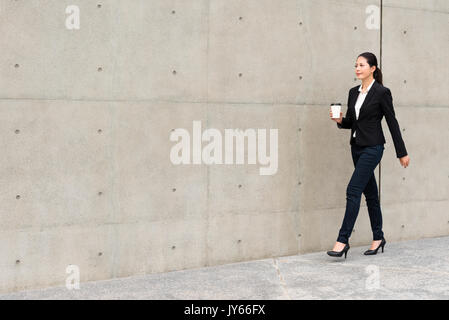 young office worker girl holding hot espresso paper cup leisurely walking on gray wall background thinking about work planning relax during lunch brea Stock Photo