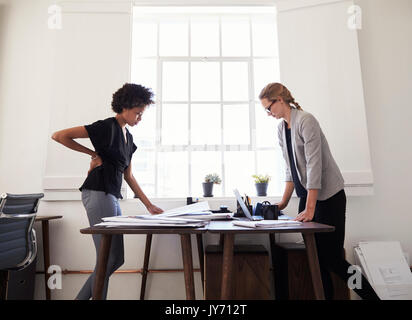 Two women stand working at opposite sides of an office desk Stock Photo
