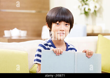 portrait of an asian little boy sitting on couch at home holding a book looking at camera smiling. Stock Photo