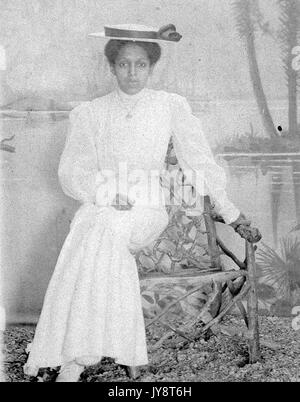 Full length portrait of African American woman wearing a hat and sitting on a handmade chair in a photographic studio, painted backdrop in the background shows a lake scene, 1915. Stock Photo