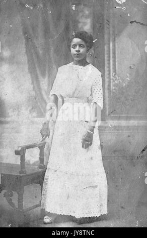 Full length portrait of young African American woman, identified as Miss Redd, wearing a white dress and standing in front of a painted backdrop, with one finger resting on the arm of a bench, 1944. Stock Photo