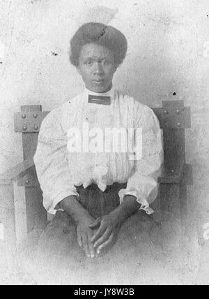 Waist up portrait of seated African American woman, wearing a white blouse and checkered dress, with a serious facial expression, hands rested on lap, 1920. Stock Photo