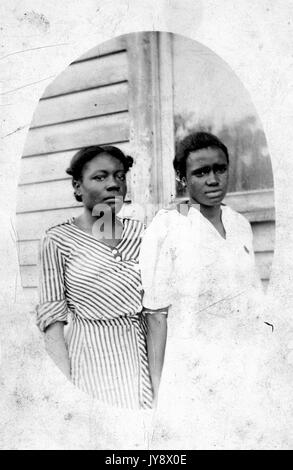 Three quarter length oval portrait of two young African American women, standing in front of a house, the one on the left in a striped dress, the other in a white dress, both with strained facial expressions, 1920. Stock Photo