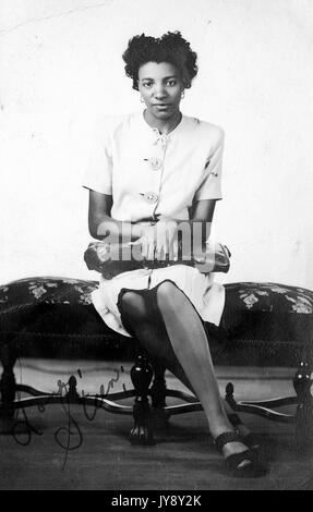 Full portrait, seated African American woman on floral print bench, wearing knee-length dress, bag or purse in lap, neutral facial expression, 1920. Stock Photo